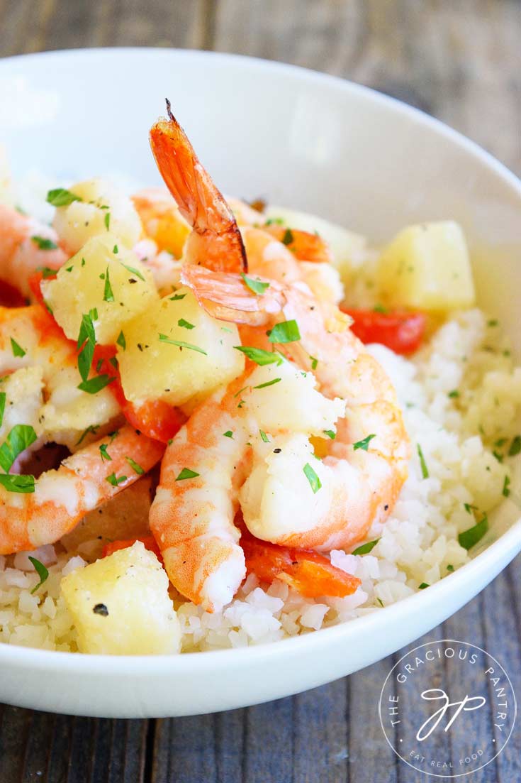 Close up shot of this clean eating sheet pan pineapple shrimp recipe showing large shrimp and chunks of pineapple on a bed of riced cauliflower and topped with chopped, green garnish. Rice is an option as well.