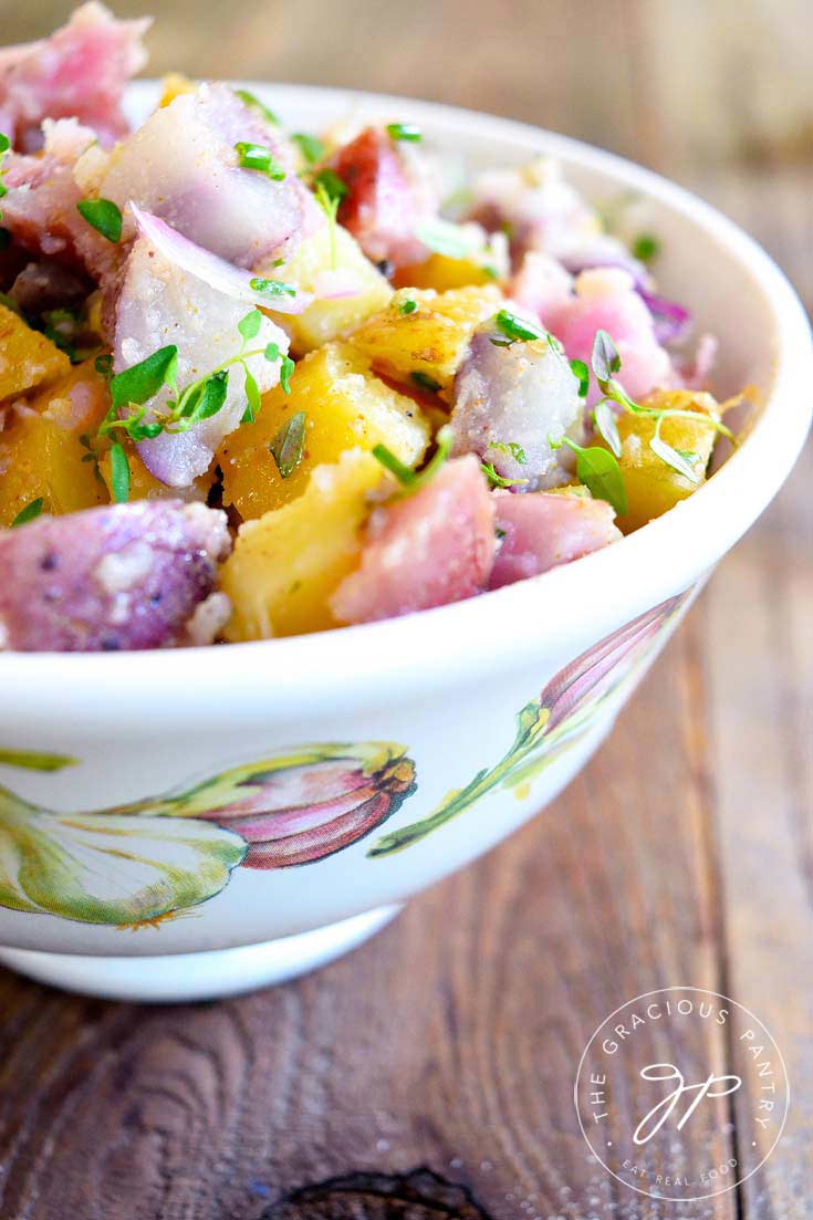 A decorative bowl sits on a wooden table top filled with this Clean Eating No Mayo Potato Salad. You can see chunks of various colored potatoes that have been sprinkled with fresh herbs.