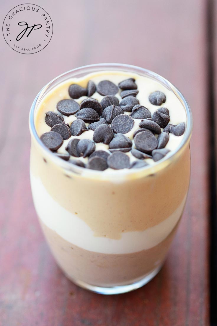 A single Clean Eating Chocolate Peanut Butter Parfait sits on a wood table. The up close image shows the parfaits layers throughs he clean glass container. Light brown, white, then yellow on top with chocolate chips sprinkled over the top layer for garnish.