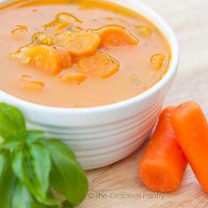 A bowl of this Cherry Tomato Soup sits served on a table with a few carrots and fresh basil next to it.