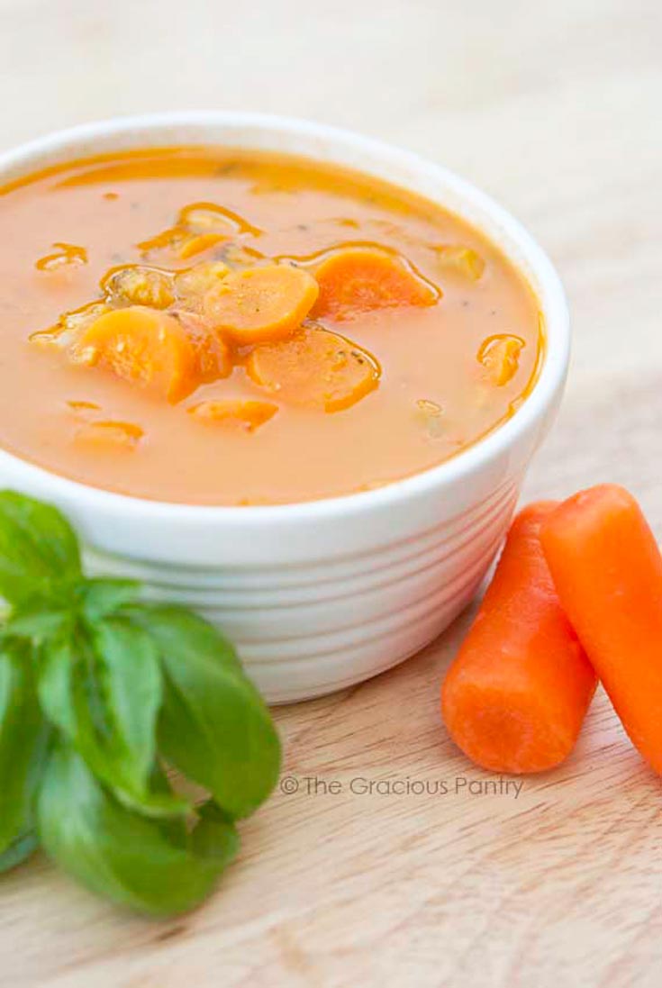 A bowl of this Cherry Tomato Soup sits served on a table with a few carrots and fresh basil next to it.