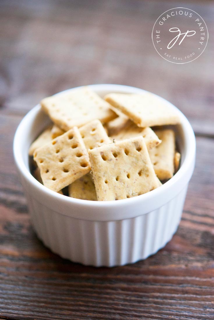 A small white bowl sits on a wooden table top, filled to the top with these square, Clean Eating Grain Free Rosemary Crackers.