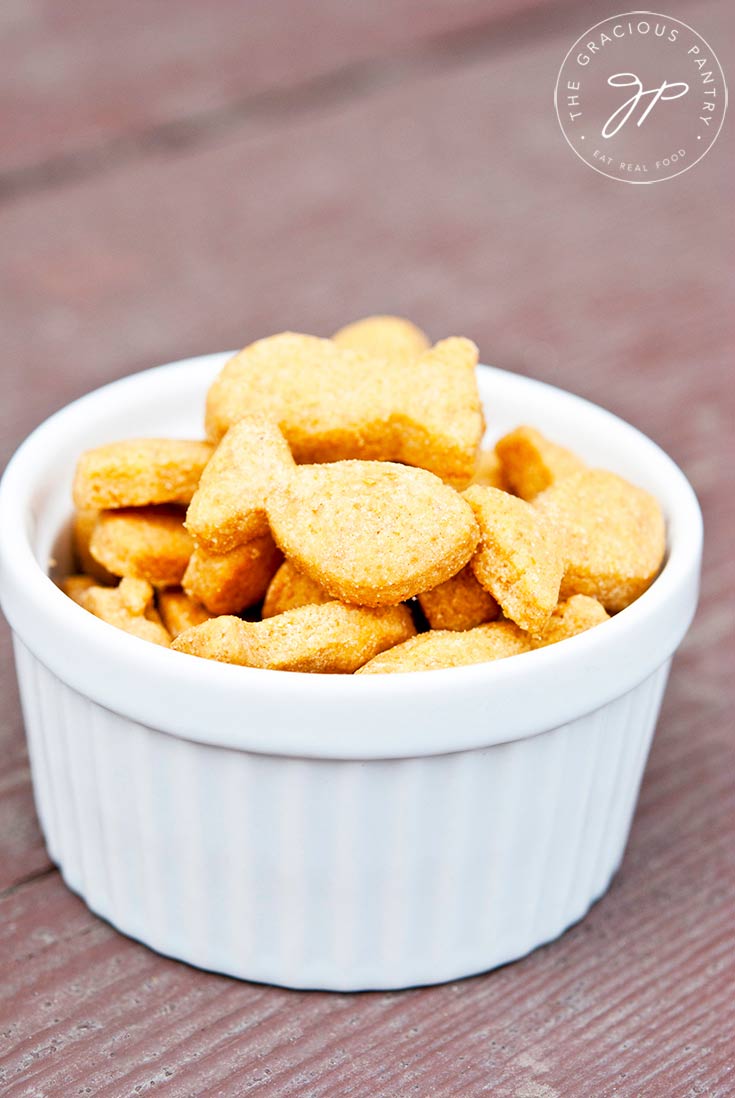 A white bowl of these Clean Eating Goldfish Crackers sits on a wooden background. The crackers are piled high and are a bright orange color.