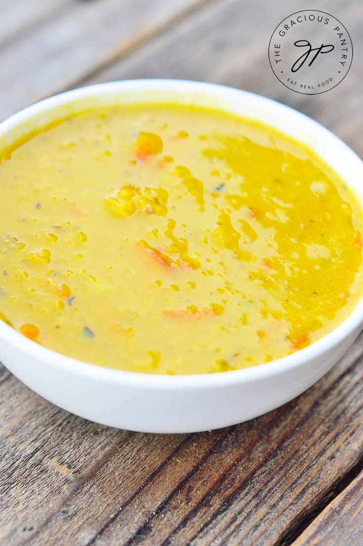 A white bowl of this Clean Eating Carrot Ginger Red Lentil Soup sits on a wooden table. You can see orange bits of carrots throughout the bright, yellow soup.
