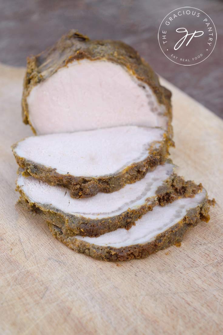 An herb crusted pork loins sits on a cutting board with several pieces sliced off of it.