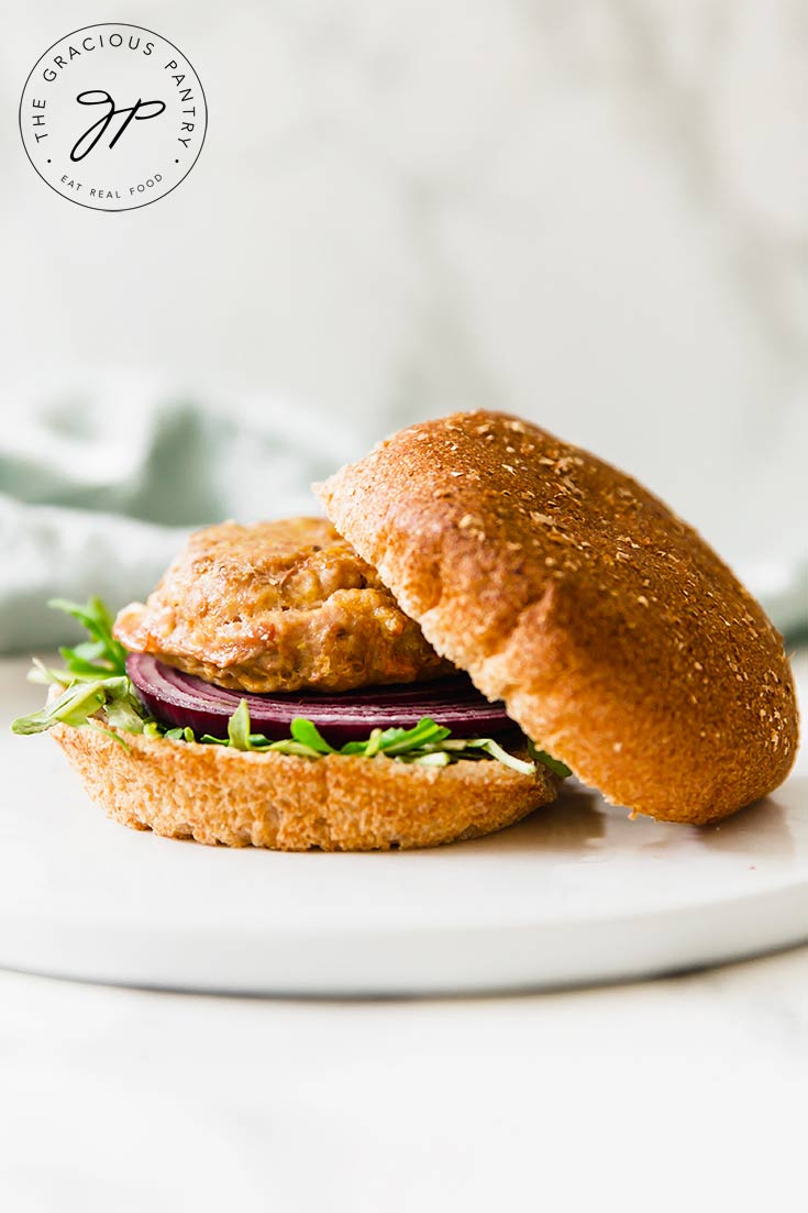 One of a batch of these Clean Eating Baked Turkey Burgers sits on a whole-grain bun, with the top bun sitting off to the side. You can see the meat patty sitting on a slice of red onion and bed of green lettuce.