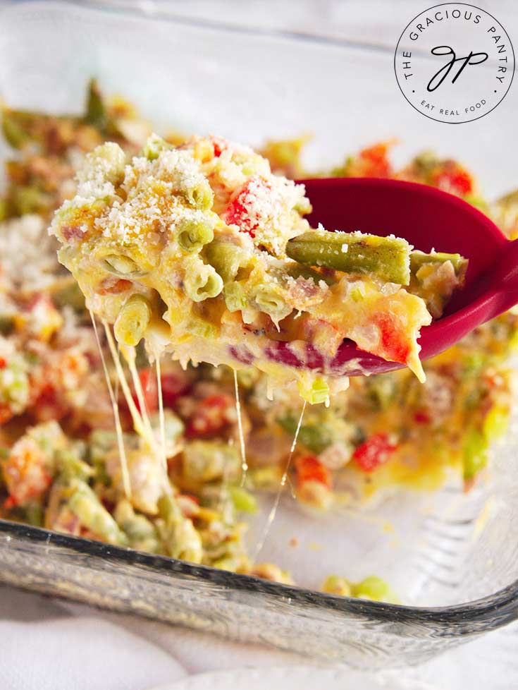 A No Noodle Tuna Casserole sits on a white background. A red serving spoons has scooped up a portion of casserole and it holding it up to the camera.