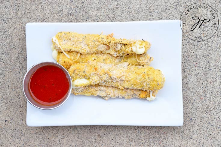 Clean Eating Air Fryer Cheese Sticks Recipe shown from overhead showing the cheese sticks stacked on a white, rectangular plate with a small dish of marinara next to them. The warm cheese is oozing out of the sticks in a few places.