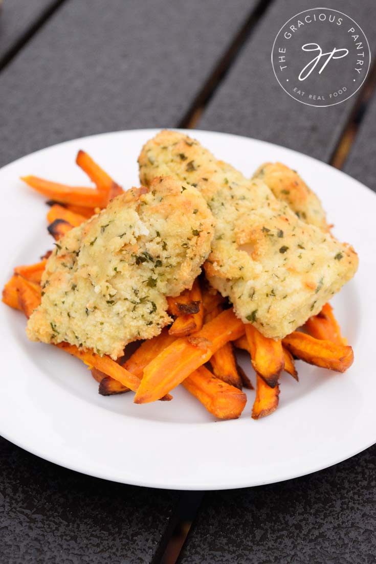 Clean Eating Air Fryer Fish N' Chips Recipe displayed on a white plate with bright orange sweet potato fries on the bottom and the golden brown, breaded fish sticks stacked on top.