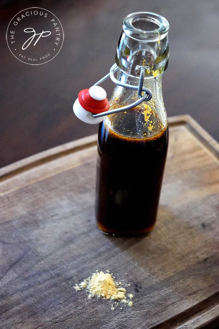 Clean Eating Teriyaki Sauce in a glass bottle with the lid open. You can see a few ginger specks on the side of the bottle from when the sauce was pour into it. There is a small sprinkle of ginger next to the bottle as well.