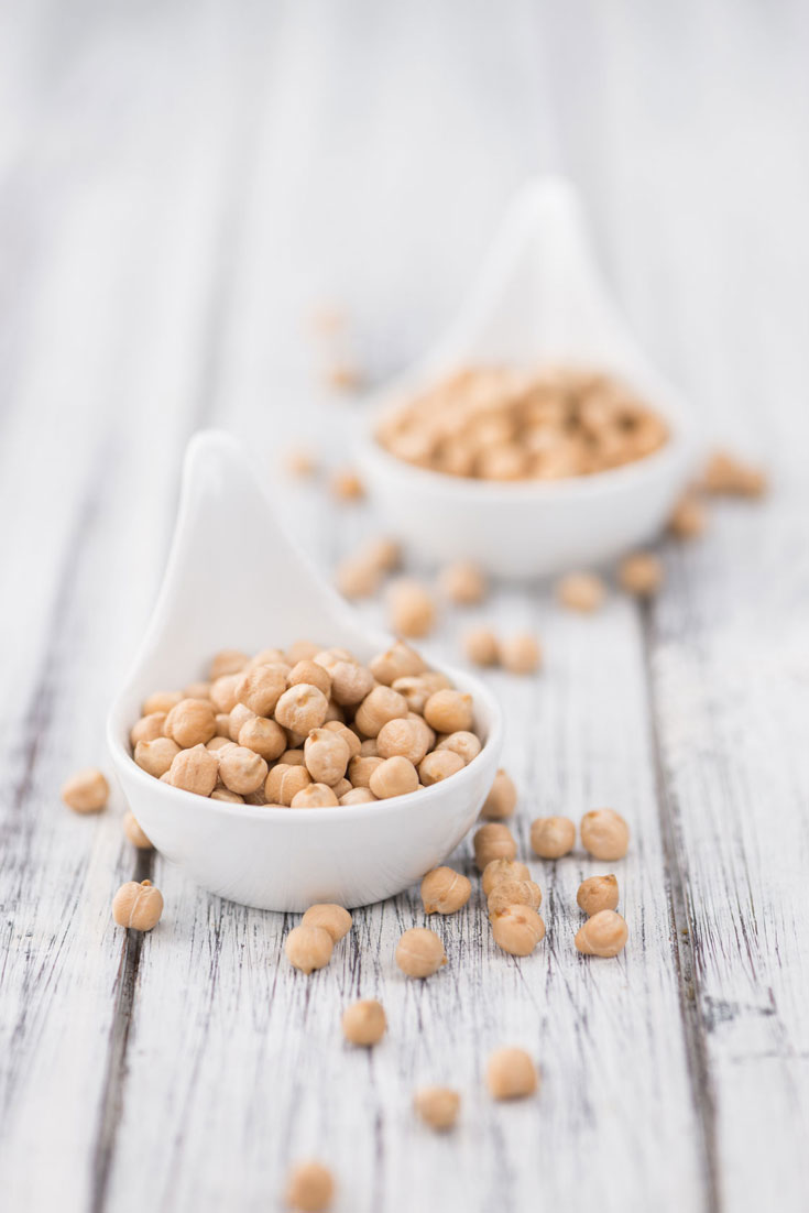 Two small, white bowls filled with chickpeas sit on a white-washed, wooden table with a few stray chickpeas scattered around the table to represent this guide on How To Cook Chickpeas In An Instant Pot