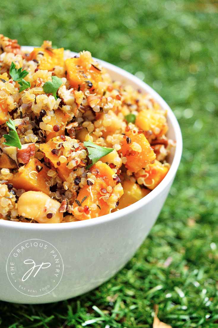 A white bowl of Clean Eating Butternut Pecan Quinoa Salad sits in green brass. You can see the grains of quinoa, bits of butternut and a sprinkle of fresh parsley over the top.