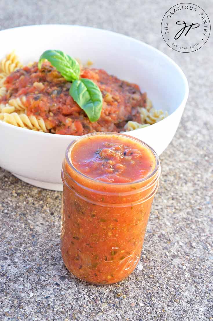 Clean Eating Instant Pot Marinara in a jar with the lid off. It's sitting next to a bowl of pasta with the sauce ladled over the top and a fresh sprig of basil sitting on top.