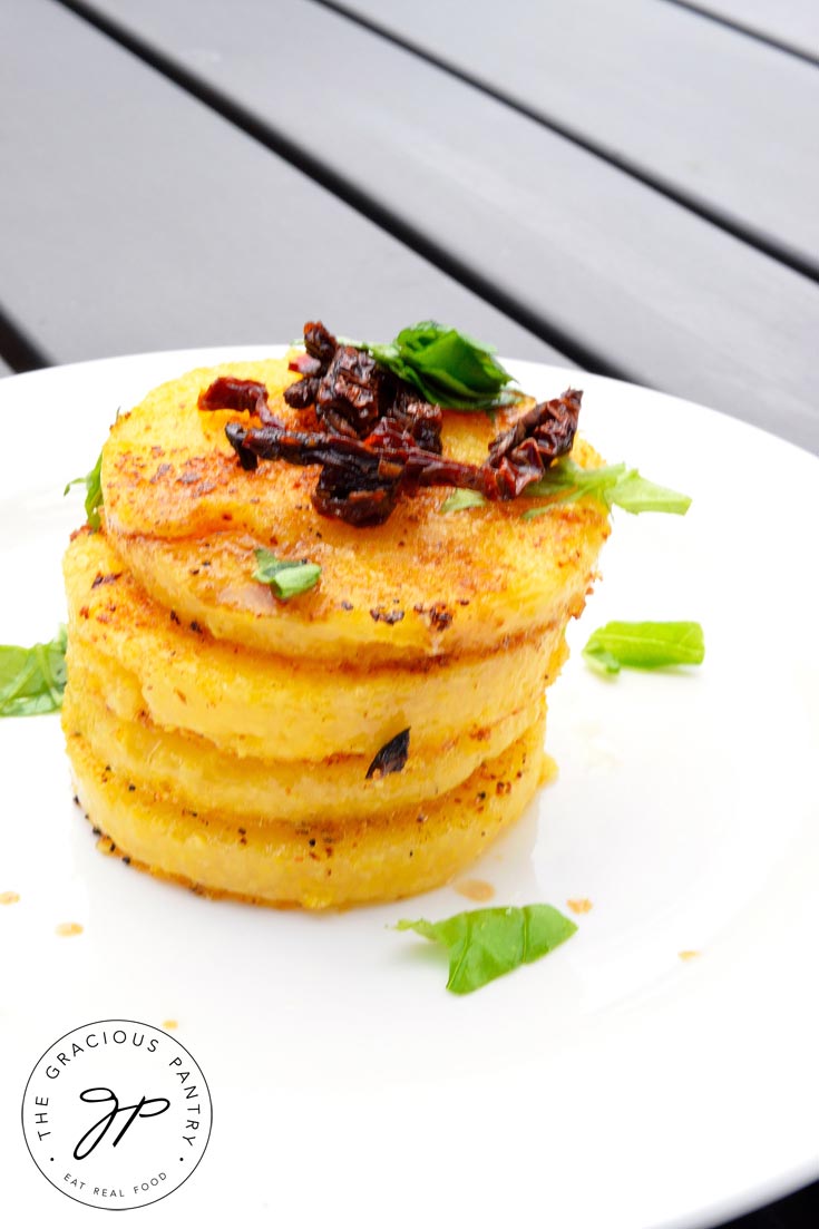 A stack of Clean Eating Italian Polenta Rounds piled high on a round, white plate. The stack is topped with chopped sun dried tomatoes and bits of fresh basil. It almost looks like a stack of cookies!