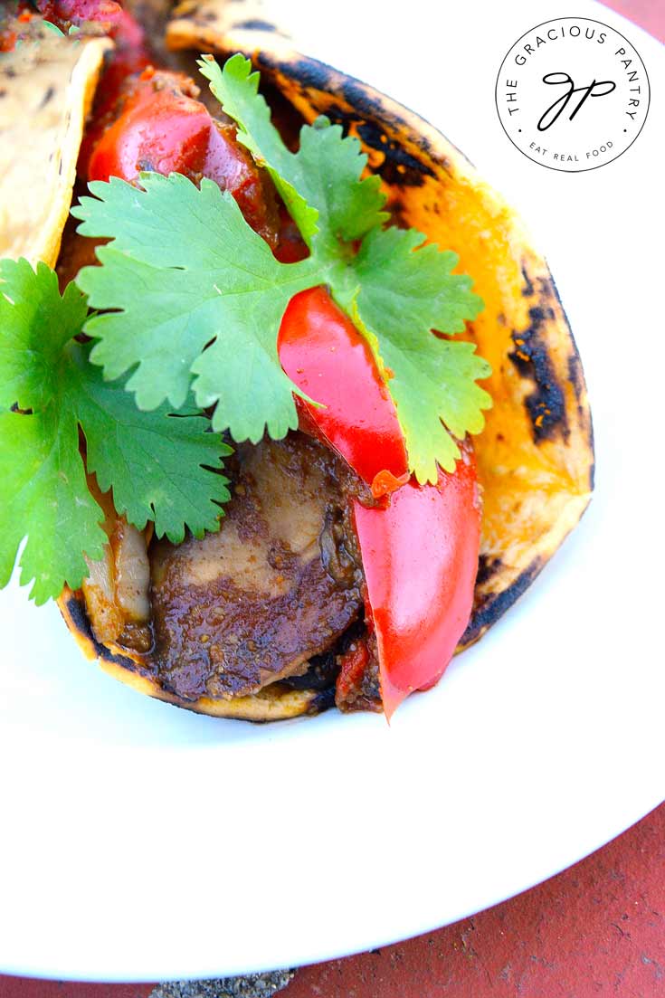 A Clean Eating Portobello Mushroom Taco sits on a white plate. You can see folded tortilla filled with cooked mushrooms and bright, red bell peppers. The taco has a tiny bit of fresh, green cilantro for garnish.