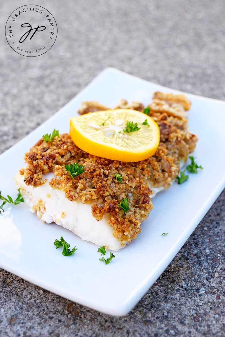 A white plate holds a fillet of Clean Eating Walnut Crusted Cod. It has a slice of lemon on top and is sprinkled with bits of fresh parsley.