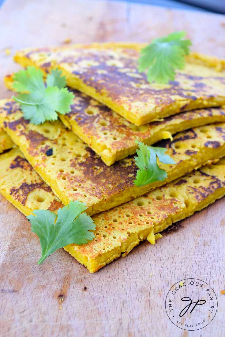 Clean Eating Chickpea Flour Flatbread cut into 4 pieces and layers on a cutting board. There are a few sprigs of fresh herbs sprinkled over the top.