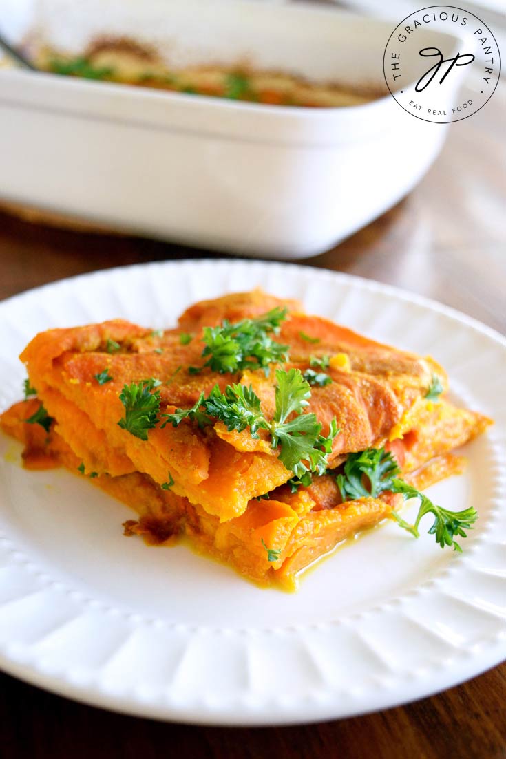 A white plate sits on a wooden table in front of a white casserole dish that this Clean Eating Layered Sweet Potato Casserole was baked in. It has a serving of the potatoes on the plate with fresh parsley sprinkled over the top for garnish.