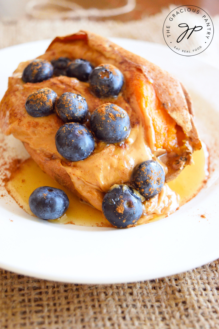 A closeup view of this Clean Eating Loaded Breakfast Sweet Potato shows just a hint of the orange flesh of the sweet potato peaking out from under a layer of creamy peanut butter topped with fresh, ripe blueberries. You can see the drizzle of maple syrup beginning to pool on the plate under the potato as well.