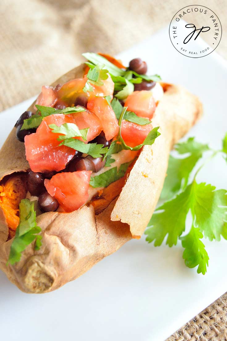 A Clean Eating Southwestern Stuffed Sweet Potato sits on a white plate with a sprig of fresh cilantro next to it. You can see the layers of ingredients starting with the sweet potato, then the hummus, black beans and finally, the tomatoes and cilantro.