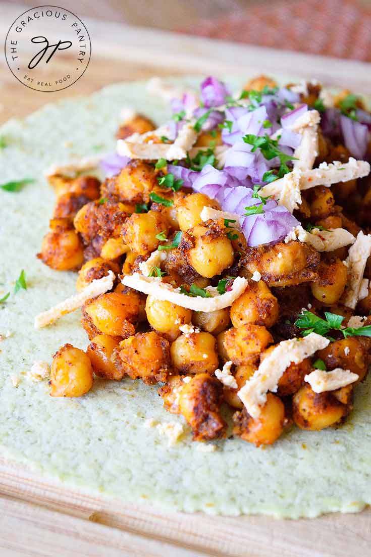 A flat tortilla sits on a cutting board layered with seasoned chickpeas, shredded cheese and chopped, purple onions in this Chickpea Tacos Recipe