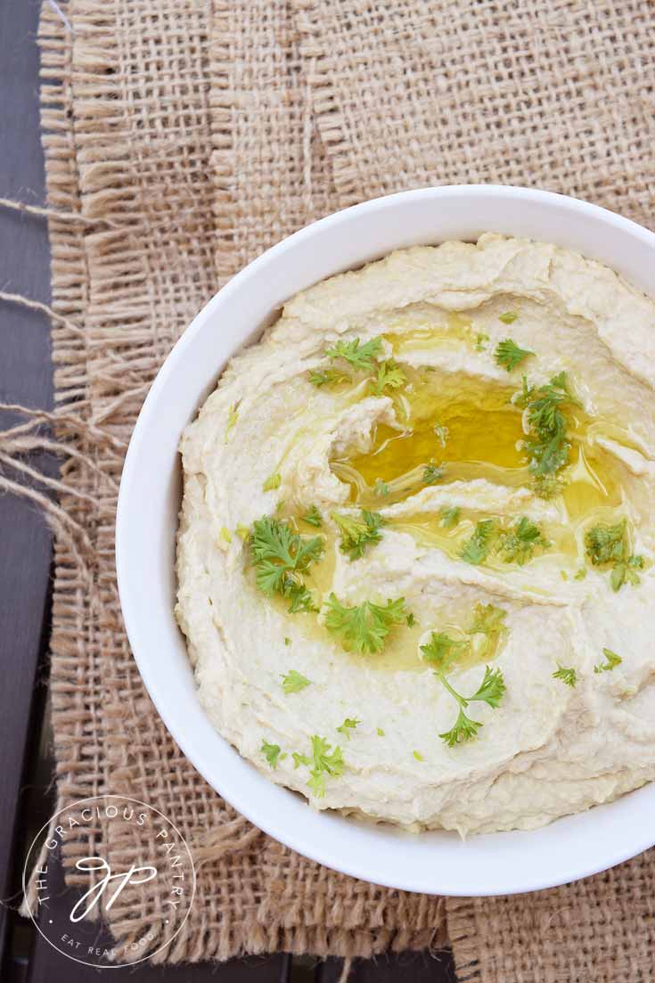 A white, round bowl sits on a piece of folded burlap on a wooden table. The view looks down into the bowl from overhead. There is olive oil drizzled over the top of the Clean Eating Artichoke Heart Hummus and there are a few sprinkles of fresh, chopped parsley over the top.