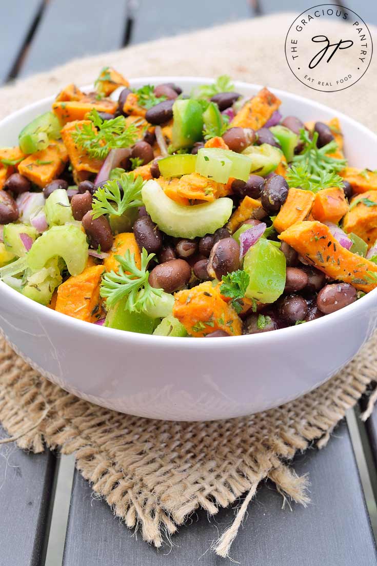 A white bowl on a dark wood table sits filled with Clean Eating Black Bean Roasted Sweet Potato Salad. You can see the beans, chunks of bright orange sweet potatoes and light green celery slices. It's topped off with chopped parsley.