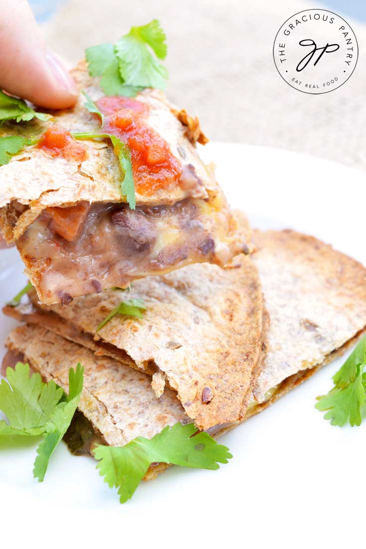 A shot of a hand holding a serving of these Clean Eating Loaded Sheet Pan Quesadillas. You can see the melted cheese oozing between the layers of the whole grain tortilla as well as a few beans peaking out. The quesadilla is topped with a small sprinkle of bright red salsa and you can see bits of fresh cilantro scattered over the top as well.