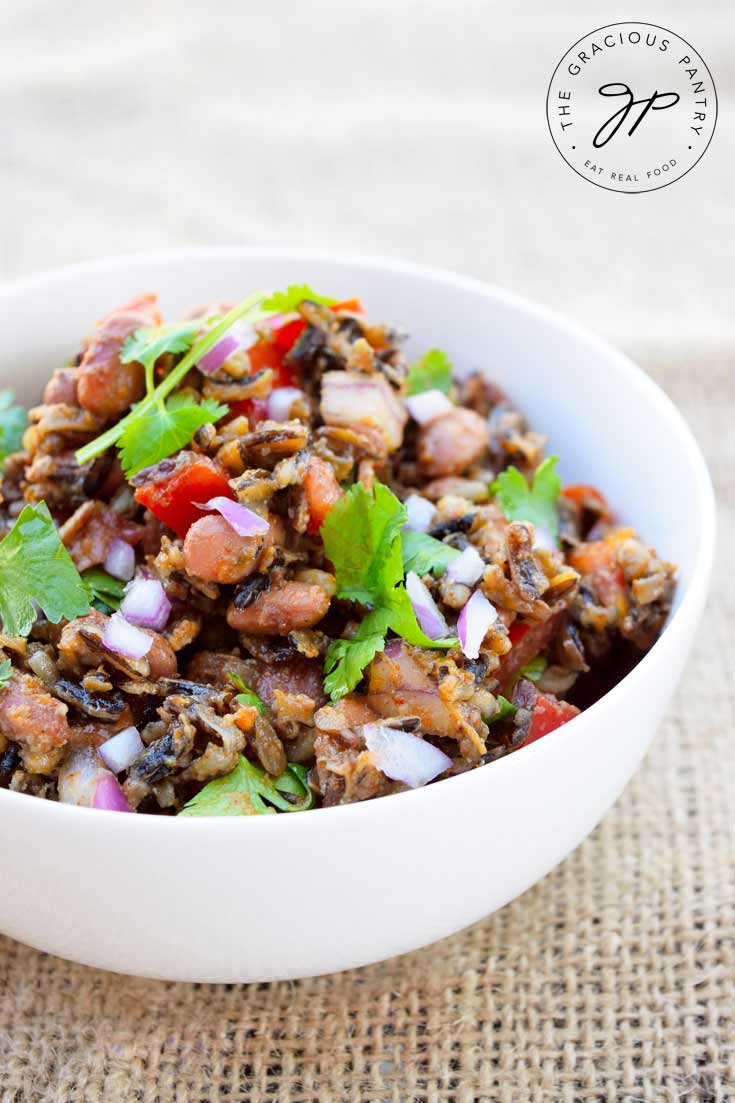 A white bowl filled with this Clean Eating Mexican Wild Rice Salad sits on a tan piece of burlap. In the bowl, you can see the rice mixed with bright red peppers, purple onions and bright green cilantro. 