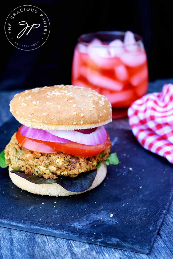 A straight on shot of one of these Clean Eating Chickpea Burgers shows the burger with a glass of red iced tea in a stemless wine glass sitting behind it and a red and white checkered napkin sitting to the right side of the burger.