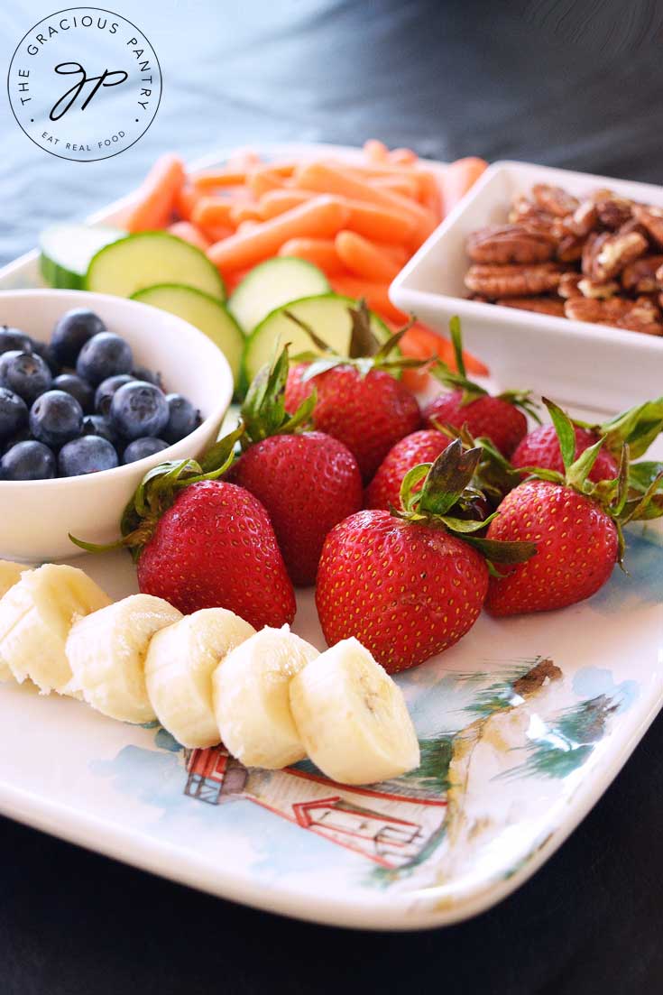 A white serving tray filled with strawberries, blueberries, bananas, pecans, baby carrots and cucumber slices to show different ideas for a Clean Eating Daily Snack Tray.