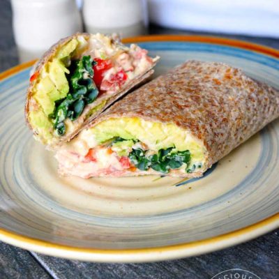 A single Kale And Hummus Wrap sitting cut, on a plate. The top half is aimed directly at the camera so you can see the layered insides of kale, hummus, tomatoes, onions and avocado.
