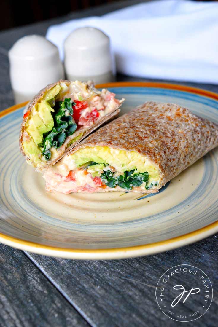 A single Kale And Hummus Wrap sitting cut, on a plate. The top half is aimed directly at the camera so you can see the layered insides of kale, hummus, tomatoes, onions and avocado.