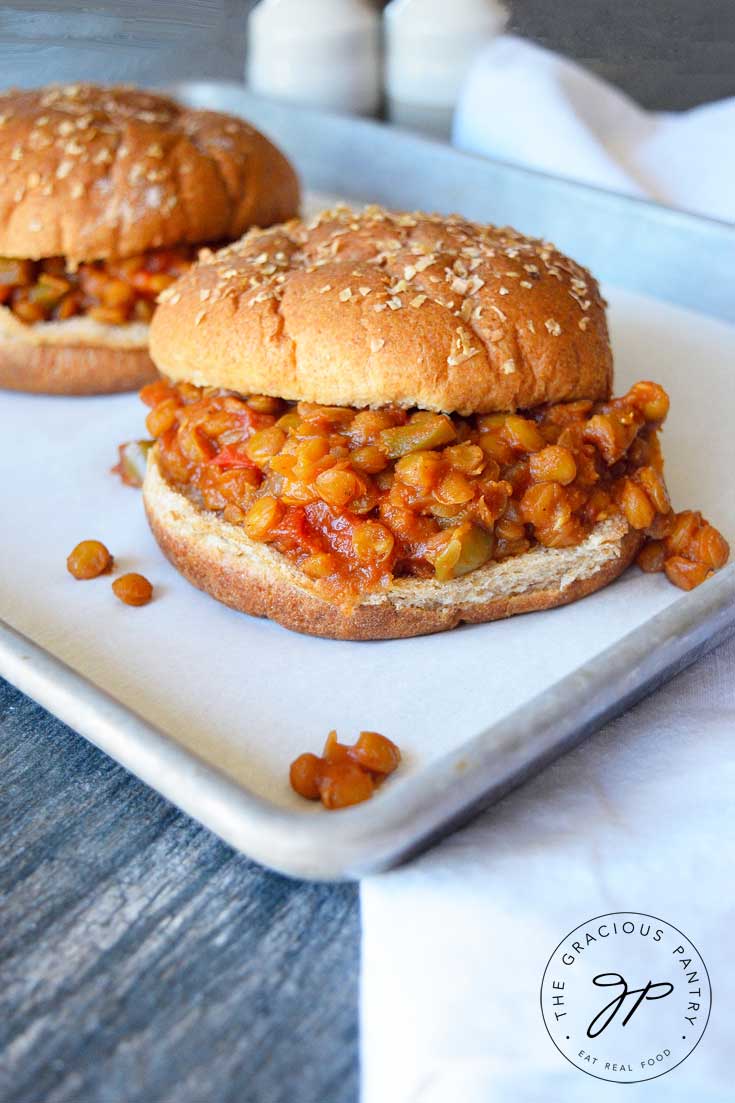 Two Clean Eating Lentil Sloppy Joes sit on a parchment-lined cookie sheet with a white towel next to it and a salt and pepper shaker in the background.