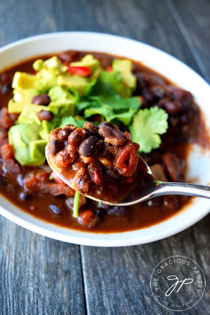 A white bowl of this Clean Eating Mexican Black Bean Chili sits in the background and a spoonful of it sits at the forefront, ready to take a bite.