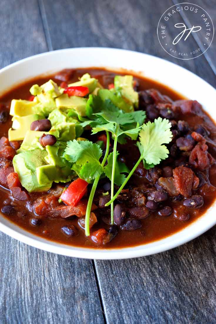 A white bowl filled with this Clean Eating Mexican Black Bean Chili and topped with fresh cilantro and chopped avocado sits on a wooden table, ready to enjoy.