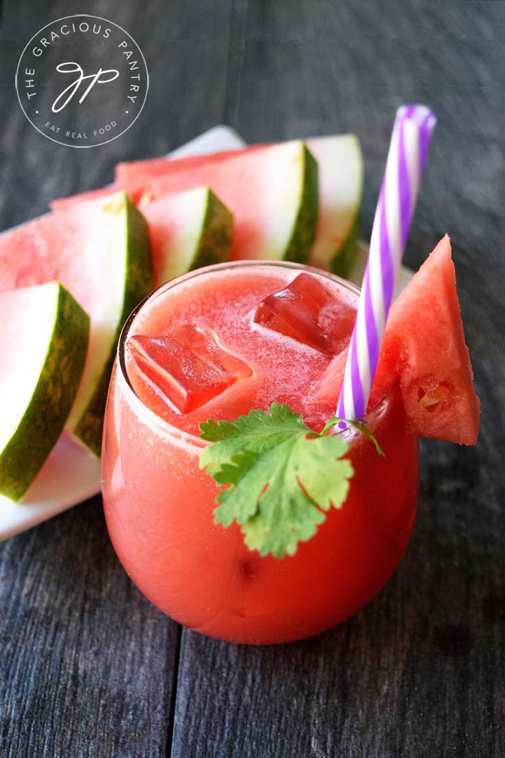 A glass of this Clean Eating Watermelon Lemonade sits next to a plate of watermelon triangles. The glass has ice in it as well as a purple-striped straw and a spring of fresh cilantro.