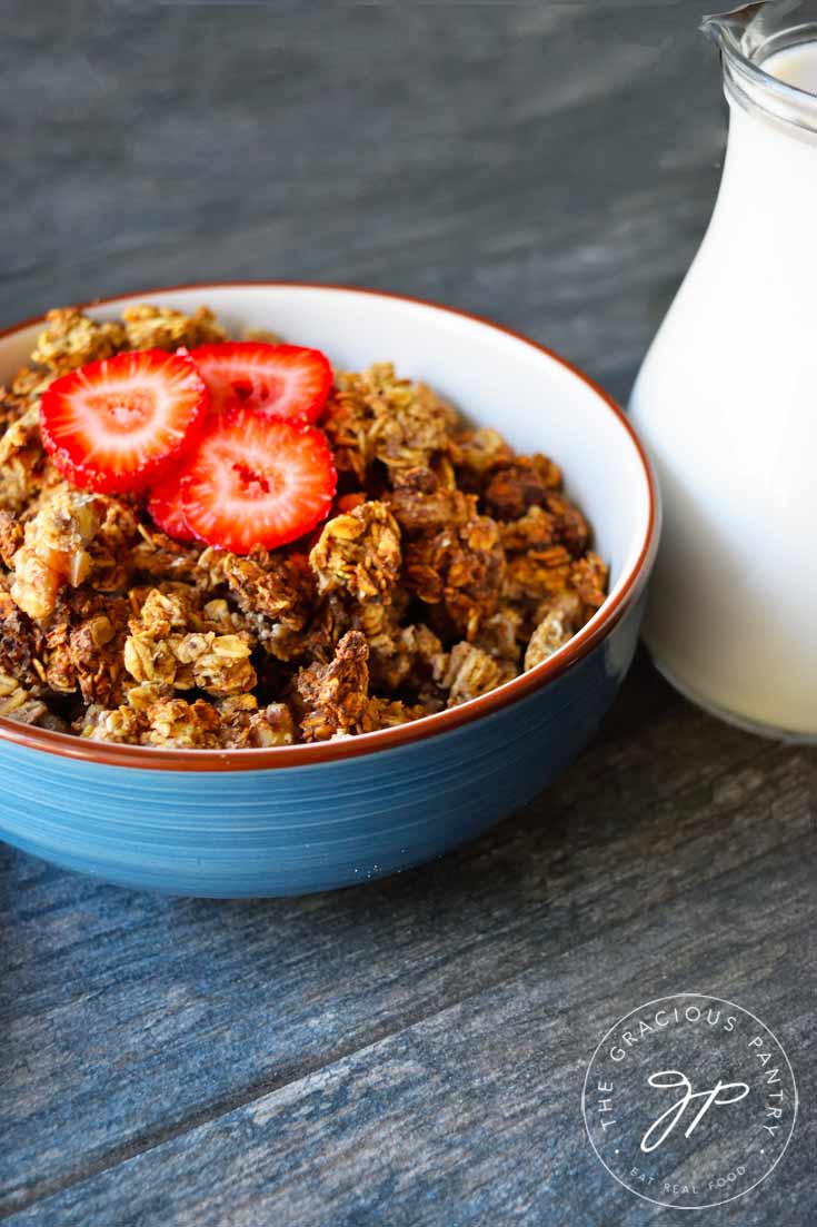 A blue bowl filled with Clean Eating Low Sugar Granola and topped with a few slices of strawberries. A pitcher of milk sits off to the side.