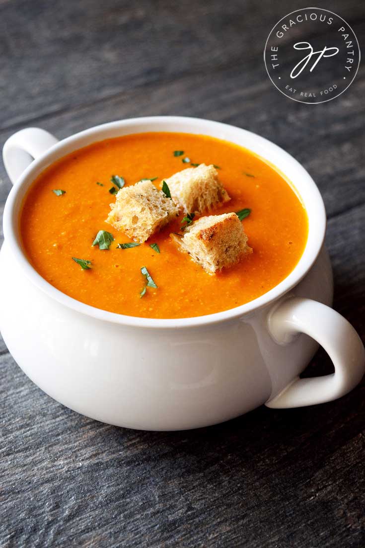 A single white bowl with handles on each side sits on a wooden surface, filled with this Clean Eating Pressure Cooker Tomato Soup. The soup has three croutons sitting in the middle and has a light sprinkle of Italian parsley over it as well.