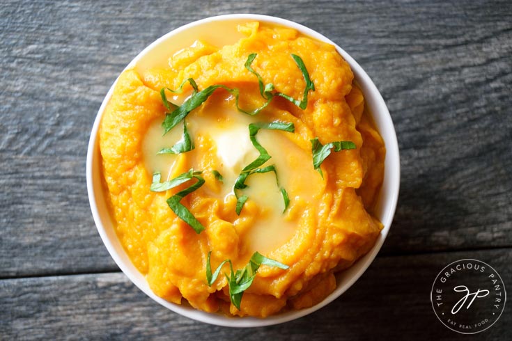 An overhead shot of this Clean Eating Instant Pot Cauliflower Sweet Potato Mash Recipe in a white bowl. The orange mash is topped with melting butter and a few sprinkles of chopped, green herbs for garnish.