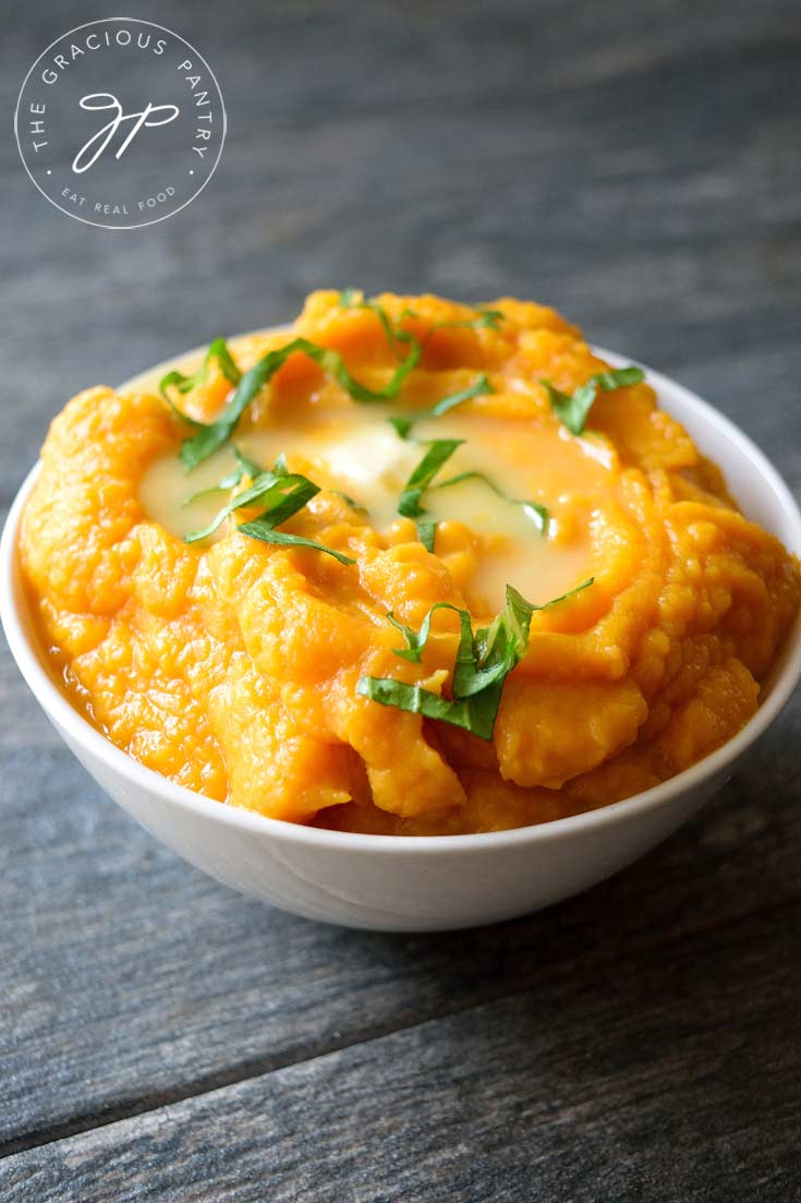 Clean Eating Instant Pot Cauliflower Sweet Potato Mash piled high in a small, white bowl with a pat of melted butter on top and a few sprinkles of fresh, green herbs for garnish.