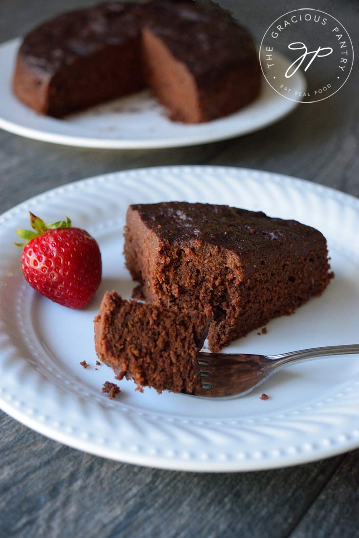 A single slice of this Instant Pot Chocolate Cake sits on a white plate with the rest of the cake in the background.