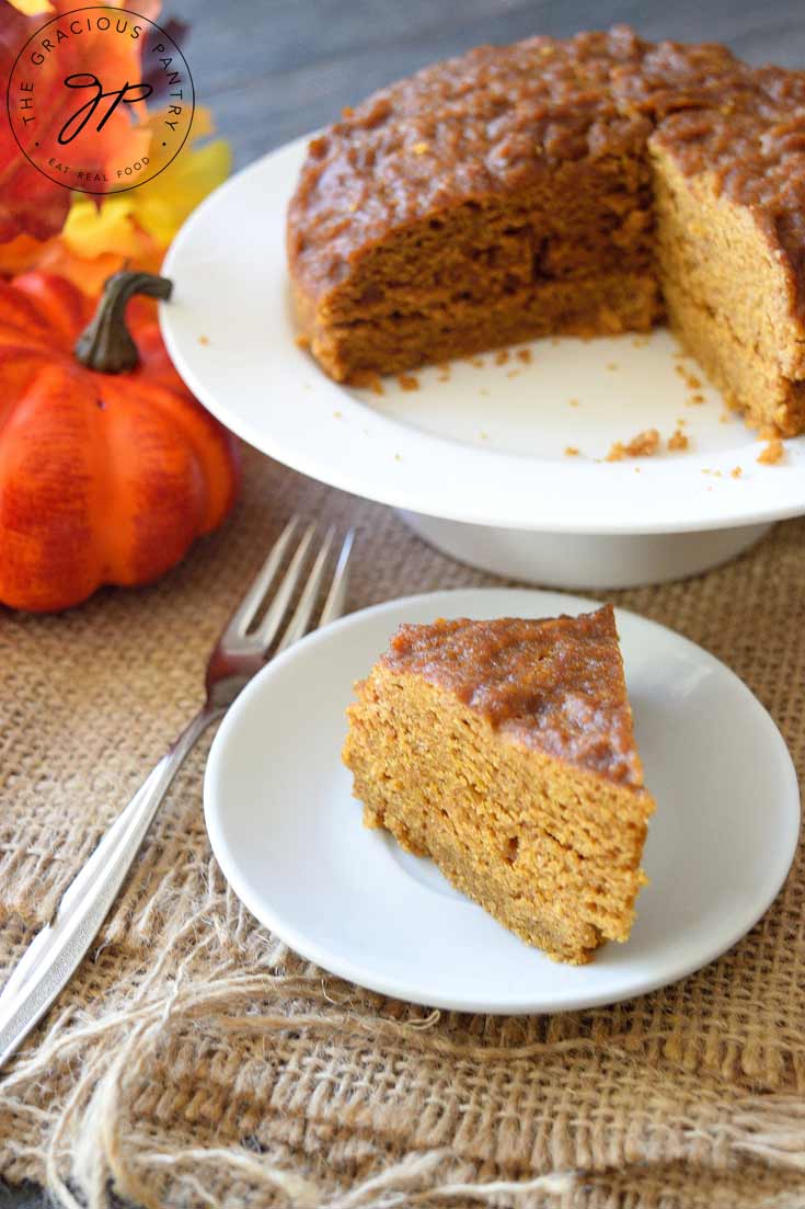 A slice of this Clean Eating Instant Pot Pumpkin Spice Cake sits in front of a cake platter with the remainder of the cake behind it. Off to the left side is a pumpkin and some autumn-colored leaves.