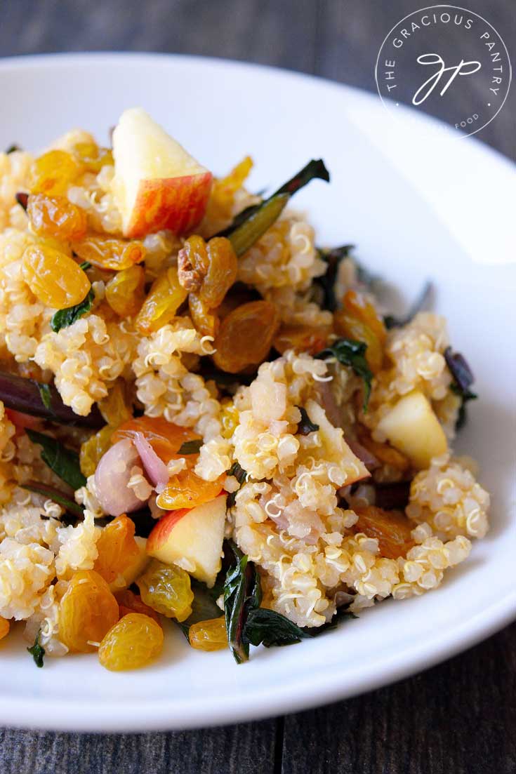 A single, white bowl filled with this Clean Eating Red Dandelion Quinoa Salad sits on a gray, wood background. You can see the bits of shallots and raisins mixed into the quinoa with the greens.