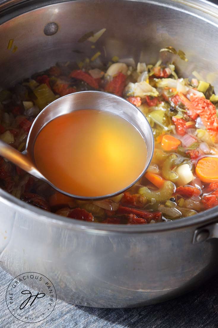 Showing this Clean Eating Vegetable Stock after it's just been made. The veggies and stock are still in the pot and ladle rests in the pot filled with golden-brown vegetable stock.