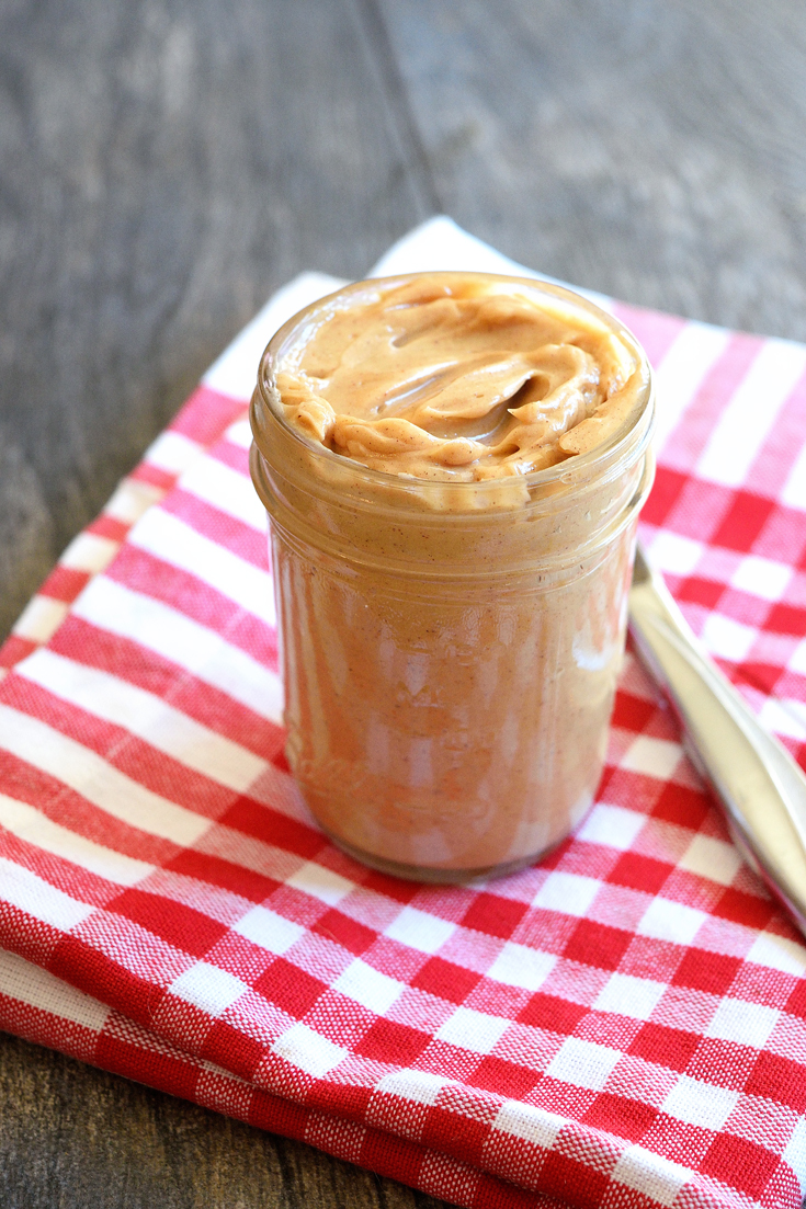 A jar of this Clean Eating Whipped Pumpkin Spice Maple Butter sits on a red and white checkered, cloth napkin. The lid is off and you can see the butter has a nice, golden brown color.