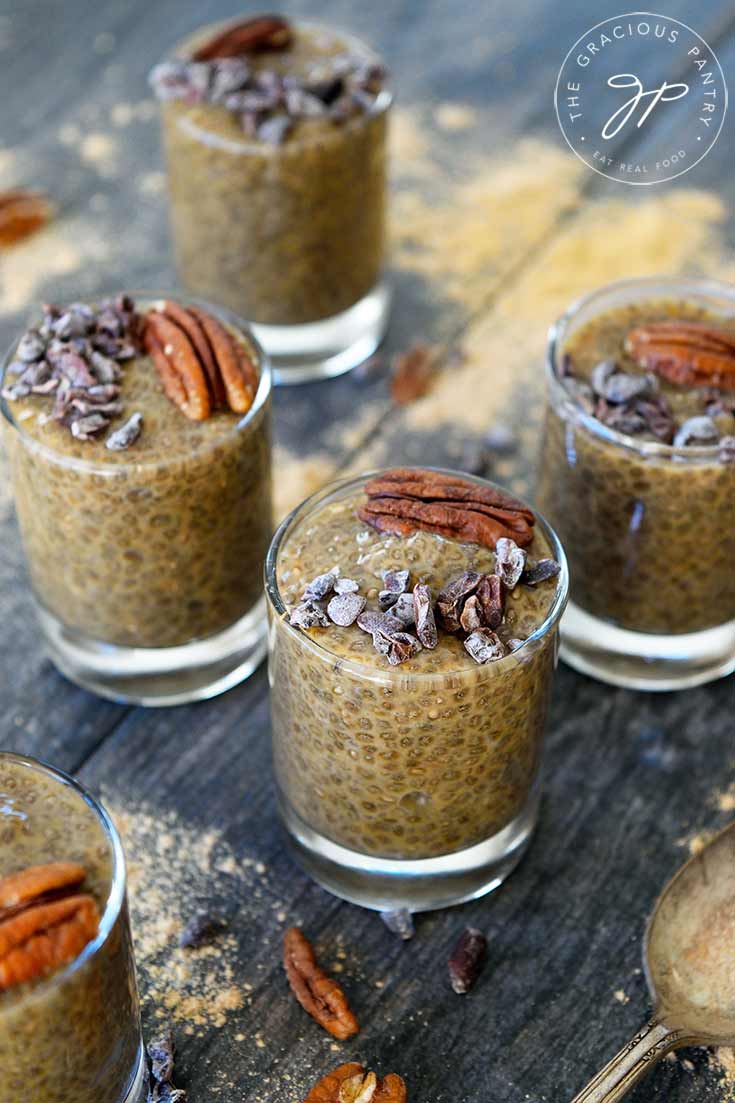 A shot of this Clean Eating Maca Chia Seed Pudding in shooter glasses, topped with a pecan half and cocoa nibs on each shooter. Maca powder is sprinkled all around the shooters and a silver spoon sits to the right of the shooters.