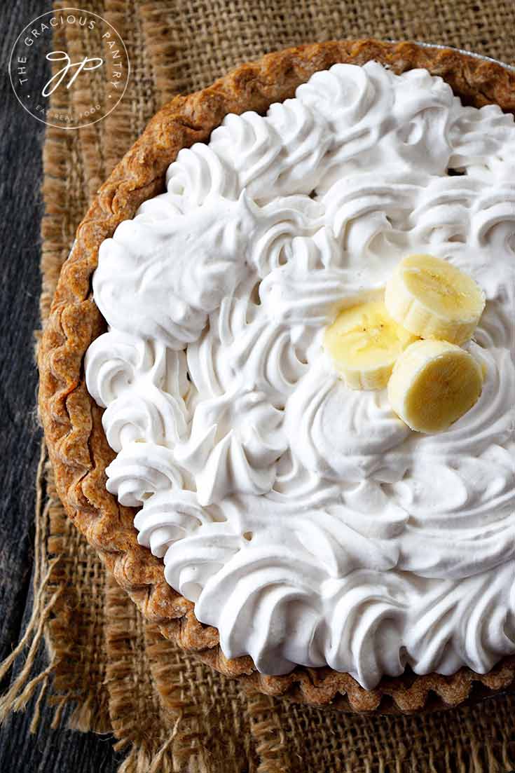A whole, clean eating banana cream pie shown from overhead. You can see the layer of swirled whipped cream on top with three slices of banana in the middle.
