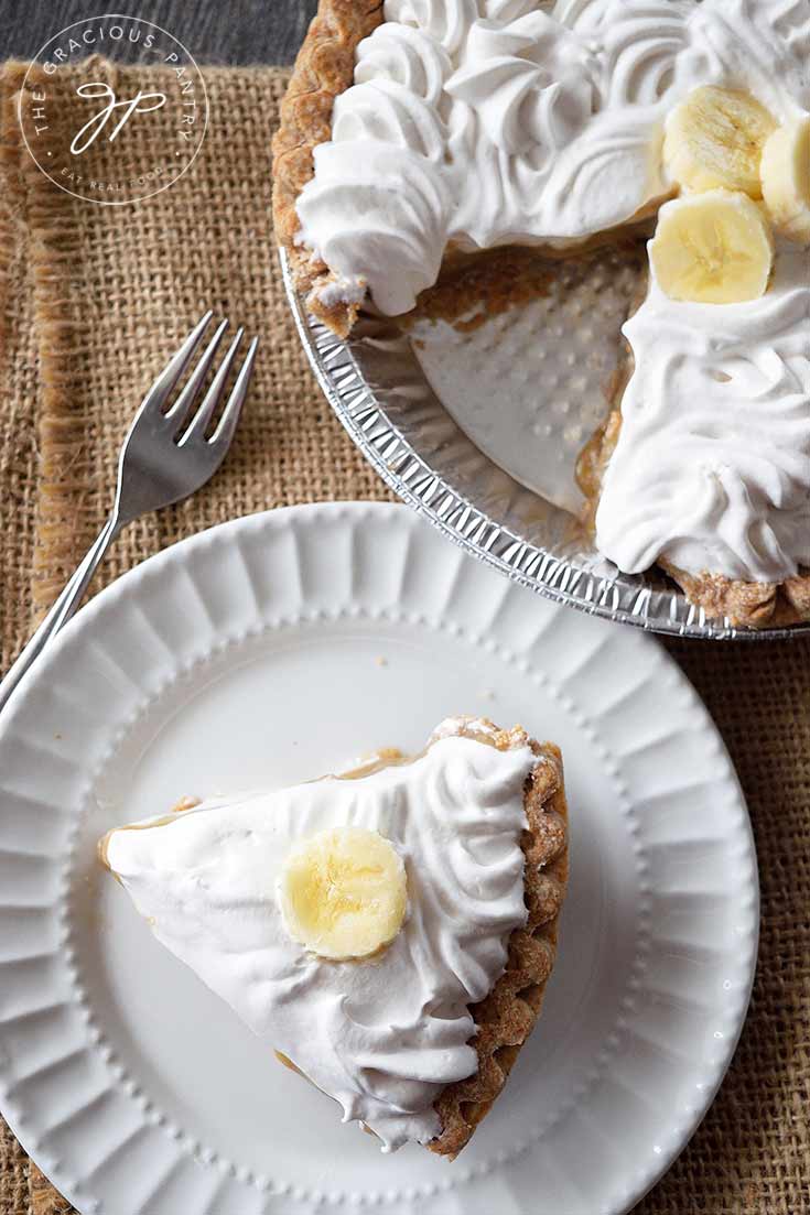 An overhead view of this clean eating banana cream pie shows the cut pie sitting next to a white plate with a single piece of pie sitting on it. A fork lays to the left of the plate.