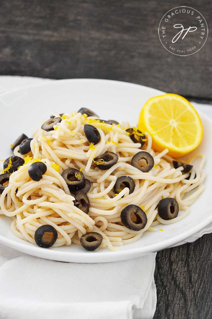A side view of this Clean Eating Olive Pasta in a white bowl with half a lemon nestled in the side of the bowl.